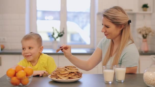 Jonge mooie moeder in blauwe shirts met plezier met haar zoon in gele overhemd op de keuken. Moeder voedt haar zoon pannenkoeken maar hij weigert. — Stockvideo