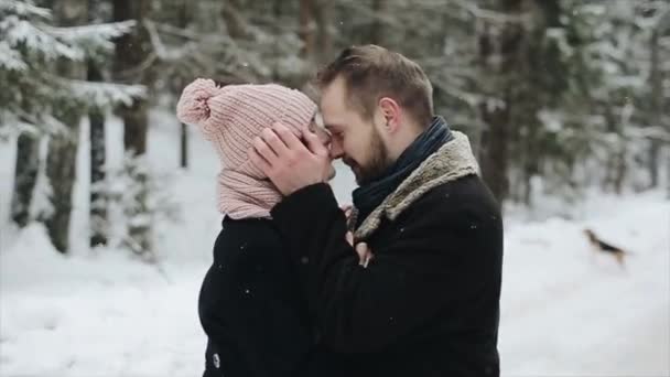 Jonge mooie Kaukasische paar zoenen onder een sneeuw in een winter forest. Paar plezier. Man trekt haar hoed in haar ogen. Slow motion. — Stockvideo