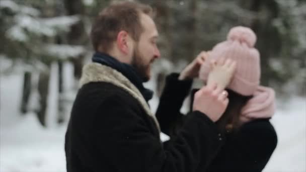 Unga vackra kaukasiska par kyssar under en snö i en vinter skog. Par har roligt. Mannen drar hennes hatt över hennes ögon. Slow motion. — Stockvideo