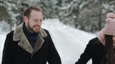 Genç çekici şiddetli kar yağışı altında bir kış ormanda yürüyüş ve sahip eğlenceli çift. Erkek ve kadın pembe bir kucaklama ve tutan eller toghether şapka. Mutlu aile kış orman içinde. Ağır çekim.