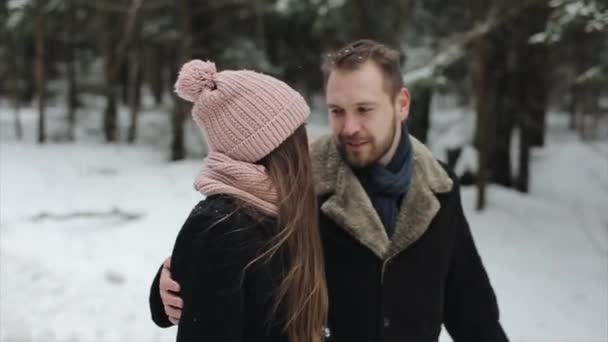 Joven pareja atractiva caminando y divirtiéndose en un bosque de invierno bajo la nieve pesada. Hombre y mujer con sombrero rosa abrazándose y tomándose de las manos. Familia feliz en el bosque de invierno. Movimiento lento . — Vídeos de Stock
