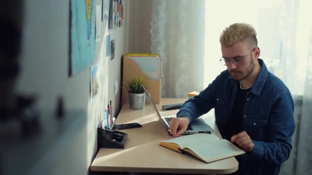 Fröhlicher junger männlicher Student mit Brille und gelben Haaren, der zu Hause arbeitet. Schreibtisch. Professionelle Home-Freelancer blättern Video auf Notebook und erstellen Artikel im Tagebuch. — Stockvideo