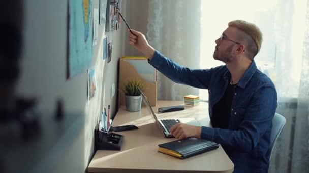 Joven estudiante alegre con gafas y pelo amarillo trabajando en casa. Escritorio. Experto freelancer casero toma notas y crea un artículo en el bloc de notas . — Vídeo de stock