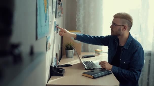 Fröhlicher junger männlicher Student mit Brille und gelben Haaren, der zu Hause arbeitet. Schreibtisch. Der gelernte Freiberufler macht sich Notizen und erstellt Artikel im Notizblock. Arbeit in der Wohnung — Stockvideo