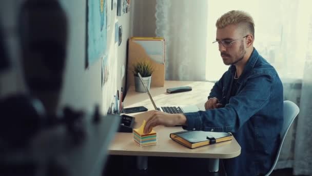年轻的有魅力的男人眼镜和黄色的头发使用笔记本电脑在家里的办公室, 并在纸贴纸上做笔记。家庭自由职业者在办公桌上的问题。时尚成功的人在办公桌前工作. — 图库视频影像