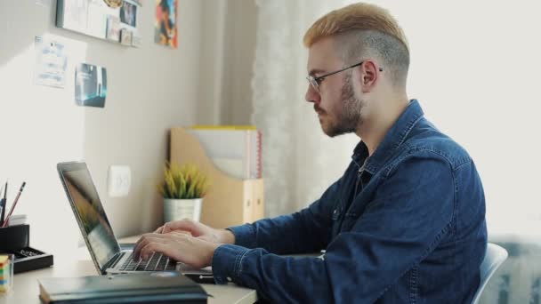 Νέους ελκυστικές επιχειρηματίας φορώντας γυαλιά με κίτρινα μαλλιά εργασία στο σπίτι. Μιλώντας στο τηλέφωνο κάθεται στο γραφείο του με το laptop. Παίρνει κακές ειδήσεις. Απογοητευμένος σαν θυμωμένος σπίτι freelancer. — Αρχείο Βίντεο