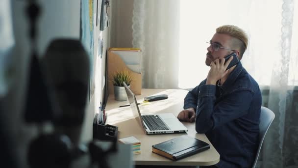 Young attractive businessman wearing glasses with yellow hair working at home. Talking on the phone sitting at the desk with laptop. Gets bad news. Disappointed san angry home freelancer. — Stock Video