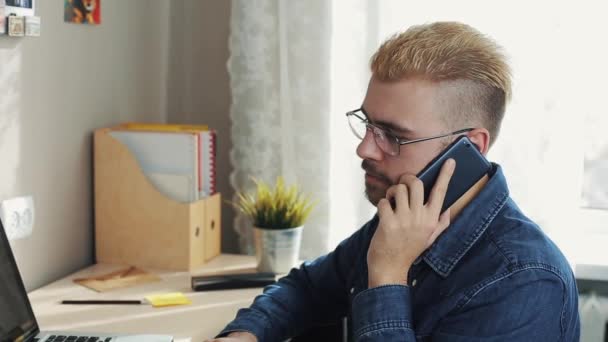 Potrait de joven y atractivo hombre de negocios con gafas de pelo amarillo hablando por teléfono. Sentado en el escritorio con portátil . — Vídeos de Stock