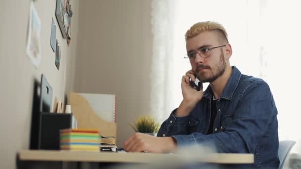 Potrait de joven y atractivo hombre de negocios con gafas de pelo amarillo hablando por teléfono. Sentado en el escritorio con portátil . — Vídeo de stock