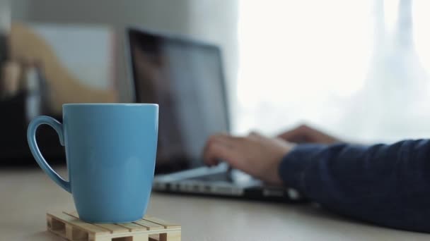 Primo piano di tazza di tè o caffè contro le mani maschili digitando su un computer portatile sullo sfondo. Libero professionista che lavora sulla scrivania . — Video Stock