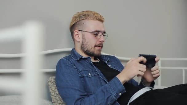Životní styl střílet atraktivní millenial mladíka s stylový účes, hraní her na smartphonu v bílé posteli. — Stock video