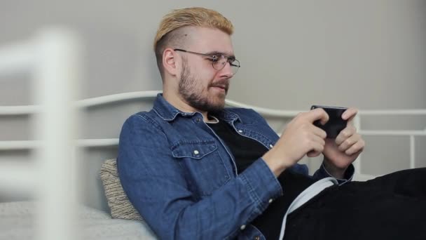 Levensstijl schieten van aantrekkelijke millenial jongeman met stijlvolle kapsel speelspel op smartphone in witte bed. — Stockvideo