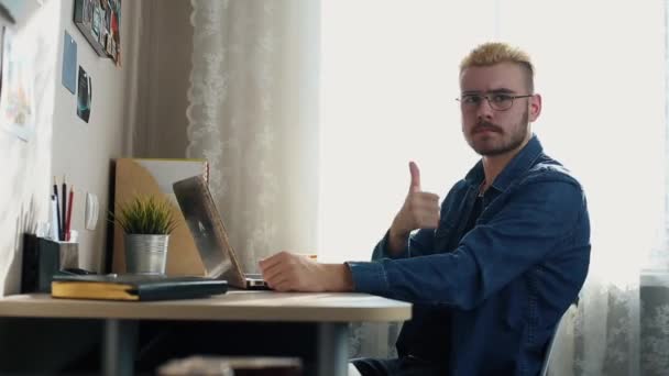 Retrato de un joven guapo con gafas de pelo amarillo que muestra el pulgar en la cámara. Home Freelancer trabajando en casa. Usando un cuaderno. Elegante hombre exitoso trabajando en el escritorio . — Vídeos de Stock