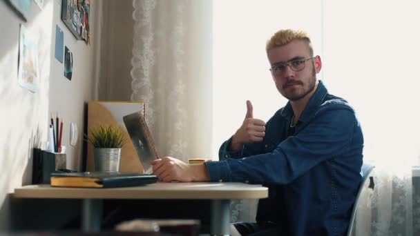 Πορτρέτο του όμορφος νεαρός άνδρας σε ποτήρια με κίτρινα μαλλιά δείχνει τον αντίχειρα στη φωτογραφική μηχανή. Αρχική Freelancer που εργάζονται στο σπίτι. Χρησιμοποιώντας ένα φορητό υπολογιστή. Κομψό επιτυχημένος άνδρας που εργάζονται στο γραφείο. — Αρχείο Βίντεο