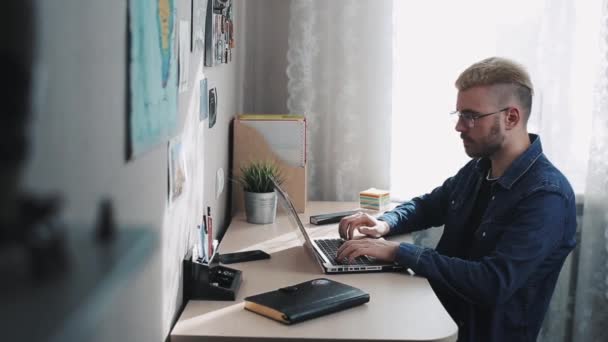 Hombre joven freelancer en casa con gafas y pelo amarillo se abre y comienza a usar Laptop. Lugar de trabajo. Oficina en casa . — Vídeo de stock