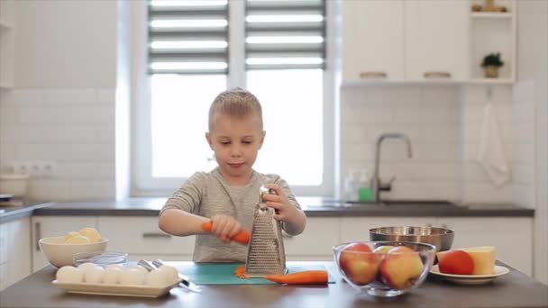 Lilla vackra kaukasiska pojke med stora blå ögon matlagning i det ljusa köket. Han är gnuggar en morot — Stockvideo