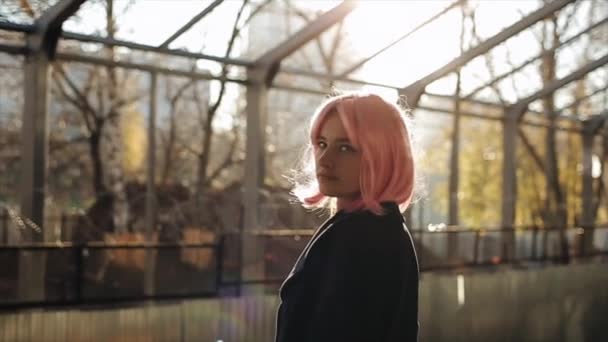 Vackra kaukasiska ung flicka med rosa hår och svart kappa stående vid järnvägsstationen. Attraktiv kvinna står mot solnedgången solen och tittar på kameran. — Stockvideo