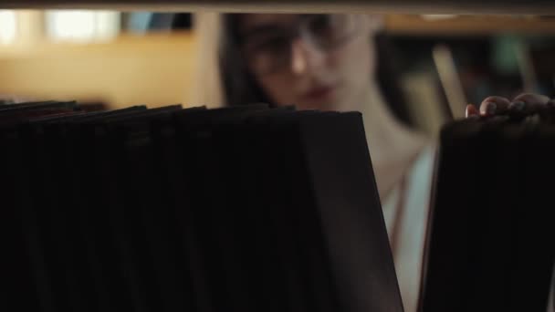 Aantrekkelijke jonge vrouw op zoek naar een boek van een boekenkast in bibliotheek — Stockvideo