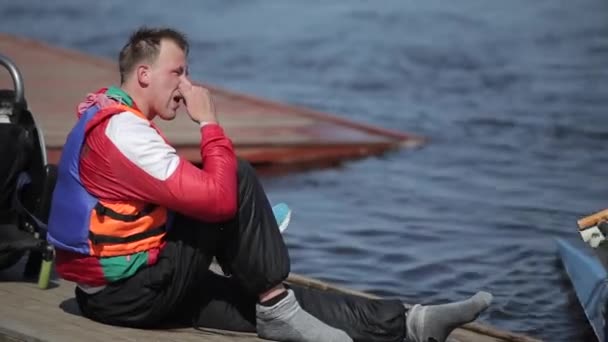 De atleet is uitgeschakeld zittend op het dok en het dragen van sneakers na training op kano. Roeien, kanoën, peddelen. Opleiding. Kajakken. — Stockvideo