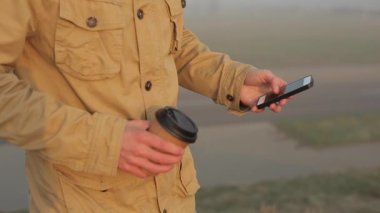 Cep telefonu kullanarak adam açık havada kapatın ve bant kaydırır. Adam kahve içiyor. Doğa