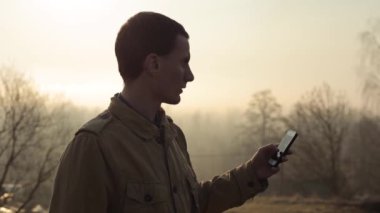 Mutlu kızıl saçlı beyaz adam arka planda ağaçlar ile bir parkta bir akıllı telefon kullanın ve bir selfie yapar. Arka planda gündoğumu veya günbatımı. Açık. Sohbet kaydırma ve internet kullanımı.