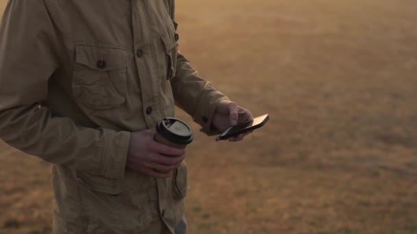 Cerrar hombre al aire libre utilizando el teléfono móvil y desplaza la cinta. Un tipo bebiendo café. el sol brilla en la pantalla — Vídeo de stock