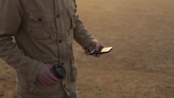 Feche o homem ao ar livre usando o telefone celular e role a fita. Um tipo a beber café. o sol brilha na tela — Vídeo de Stock