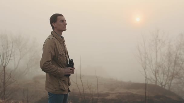 배경에 일출과 일몰을 보고 쌍안경을 들고 카키 코트를 입은 젊은 빨간 머리 남자의 클로즈업. 그의 주위에 안개. — 비디오