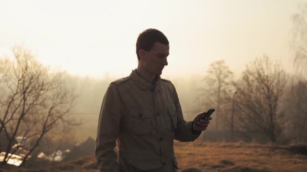 Boldog vöröshajú kaukázusi srác beszél a telefon egy parkban fákkal a háttérben. Napkelte vagy napnyugta alatt. Szabadtéri. Csevegni görgetés és használata Internet. — Stock videók