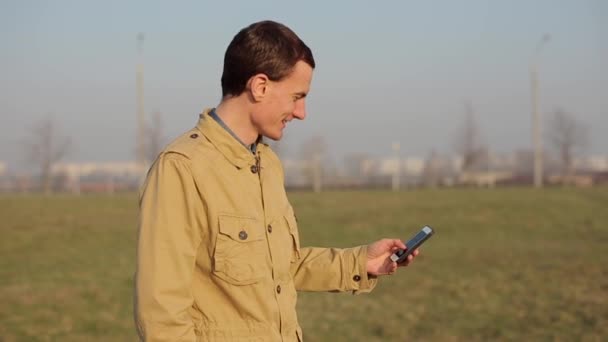 Boldog vöröshajú kaukázusi srác használja a Smart Phone egy parkban fák a háttérben, és teszi a selfie. Napkelte vagy napnyugta a háttérben. Szabadtéri. Csevegni görgetés és használata Internet. — Stock videók