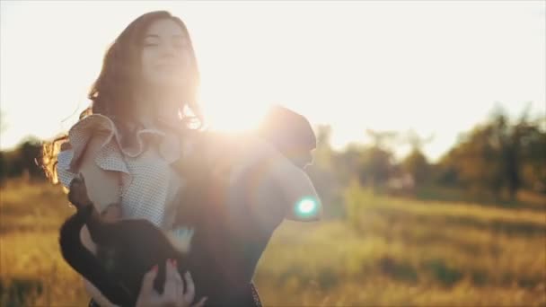 Menina bonita na moda em vestido de verão elegante e cabelo ondulado preto com cão dachshund no campo com flores à luz do sol, grama ao pôr do sol. Mulher sorri, ri e gira com o cão — Vídeo de Stock