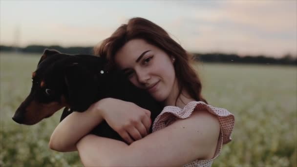 Menina bonita na moda em vestido de verão elegante e cabelo ondulado preto com cão dachshund no campo com flores à luz do sol, grama ao pôr do sol. Mulher sorri, ri e gira com o cão — Vídeo de Stock