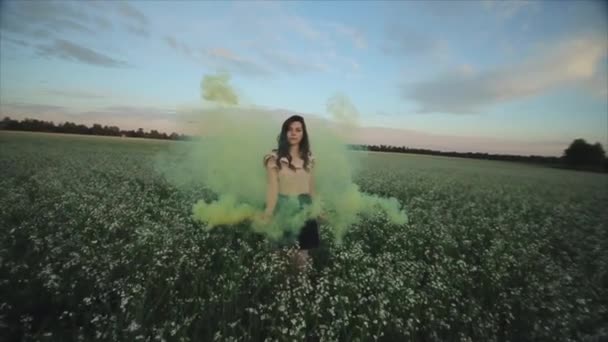 Широкий вид юной привлекательной брюнетки в юбке с зеленой дымовой бомбой на поле. Девушка веселится смеясь наслаждаясь и кружась от дыма против заката . — стоковое видео
