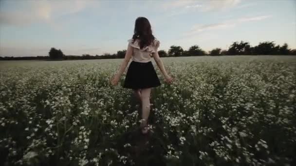 黒いスカートで波状の髪を持つかわいい若いブルネットの女の子のバックビューは、花畑を歩きます。背面図。スローモーション — ストック動画