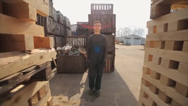 Un joven trabajador en uniforme camina entre paletas de madera en almacén de distribución al aire libre. Movimiento lento — Vídeos de Stock