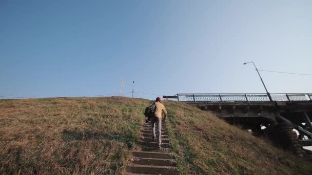 Young active man with backpack climbs the stairs on the bridge under grass mountain. — Stock Video