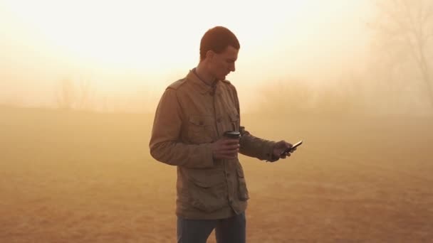 Junger Mann im Mantel trinkt seinen Morgenkaffee und benutzt und blättert Mobiltelefon mit fantastischer Aussicht bei Sonnenaufgang — Stockvideo