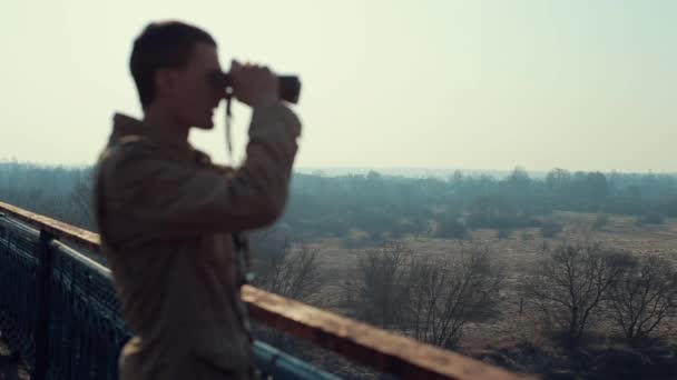 Junger Mann mit roten Haaren im khakifarbenen Mantel blickt von der Brücke durch ein Fernglas. — Stockvideo