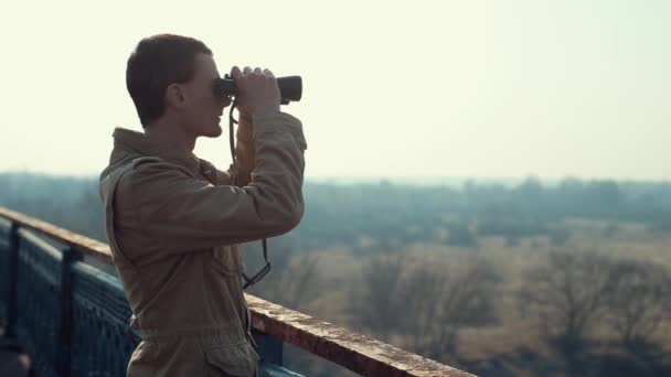 Hombre joven con pelo rojo en abrigo caqui mirando a través de los prismáticos desde el puente . — Vídeo de stock