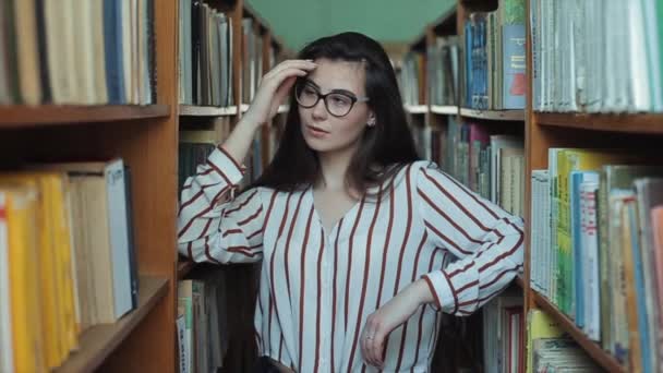 Retrato de una chica joven y hermosa en la biblioteca. Estudiante femenina estudiando entre muchos libros entre estantes . — Vídeos de Stock