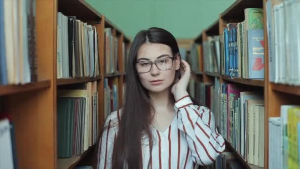 Bobruisk, Bielorrússia - 11 de abril de 2019: Retrato de uma jovem linda garota na biblioteca. Estudante do sexo feminino estudando entre lotes de livros entre prateleiras . — Vídeo de Stock