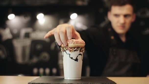Un barista poniendo una rosquilla encima de un batido en una taza de plástico y decorándolo con crema batida — Vídeos de Stock