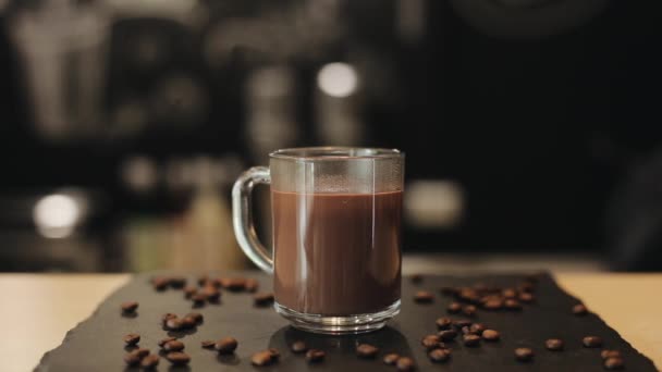 Una taza de vidrio con chocolate caliente en la mesa con granos de café. Chocolate caliente que vierte de la cuchara — Vídeos de Stock