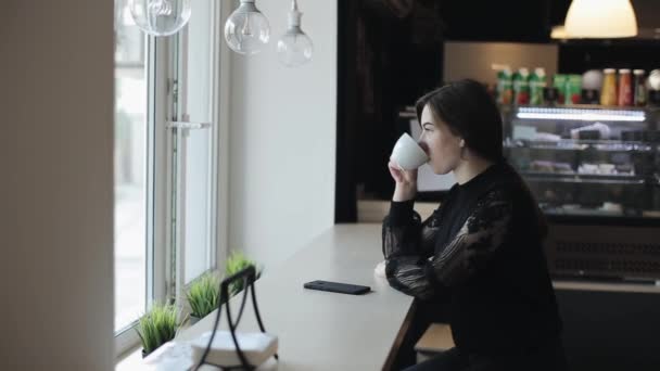 Una chica muy joven sentada en una cafetería, bebiendo una taza de té, y usando su teléfono inteligente. Una vista lateral — Vídeo de stock