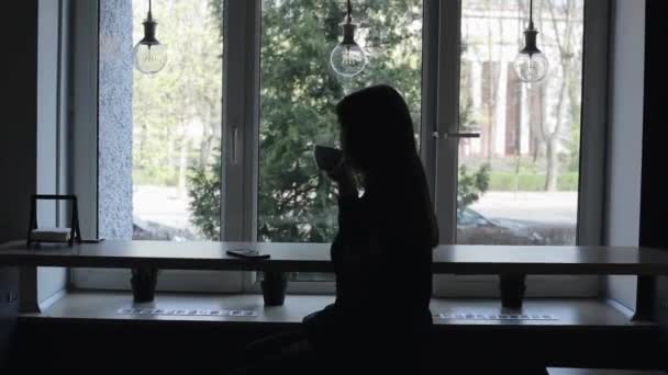 Silhouette di una bella ragazza che beve una tazza di caffè in un caffè e utilizza il suo telefono cellulare . — Video Stock
