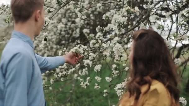 Um encantador casal jovem e gentil fica no jardim da primavera. O tipo tira as flores da árvore e dá à rapariga. Ela sorri feliz e beija sua amada. Movimento lento — Vídeo de Stock