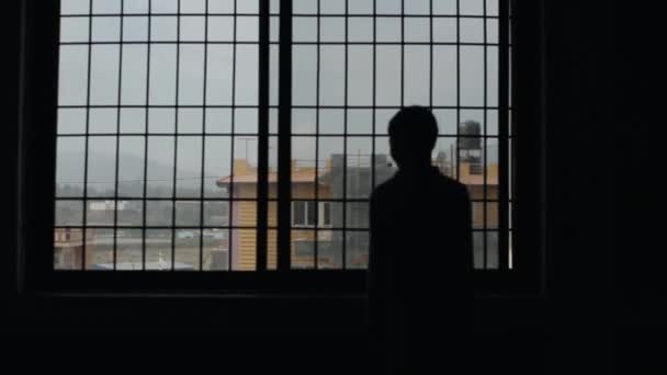Het silhouet van een piepende tienerjongen op de achtergrond van een raam met rooster in een donkere kamer. Buiten het raam de daken van huizen en bergen. De camera verandert van focus — Stockvideo