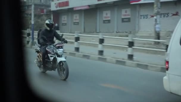 Delhi, India - 29 november 2019: Kathmandu. Nepal. Een motorrijder in een helm en een zwart jasje met een rugzak achter zijn schouders rijdt op een drukke weg door de stad. Schieten vanuit het autoraam — Stockvideo