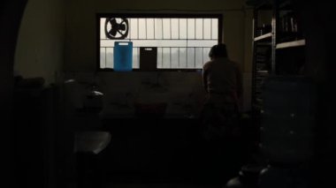 Karanlık bir mutfakta ev işleri yapan genç bir kadının silueti. Pencerenin arka planına karşı şebekesi ve manzarası olan bir kadın. Yakın plan.