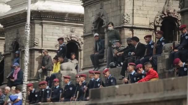 Kathmandu, Nepal - 14 de novembro de 2019: Os militares asiáticos com armas nas mãos assistem ao desempenho na rua da cidade sentados nos degraus do templo entre outros moradores e — Vídeo de Stock